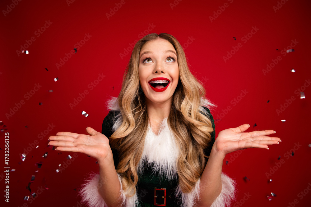 Naklejka premium Joyful woman in festive Santa style dress celebrates Christmas joy with confetti against a bold red background and sparkling moments of merry holiday spirit