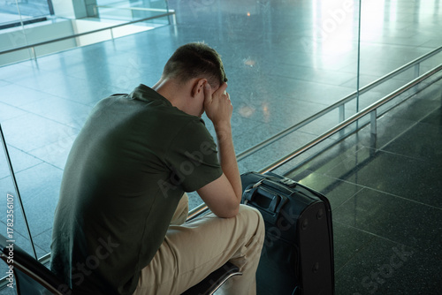 Young man with suitcase sitting in airport terminal looking upset, stressed and disappointed. Concept of flight delay, cancellation or travel problems.