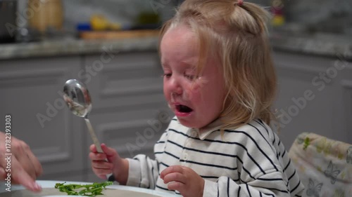 Blonde toddler in striped shirt cries intensely with raw emotion, defiantly refusing green food on plate during mealtime struggle.