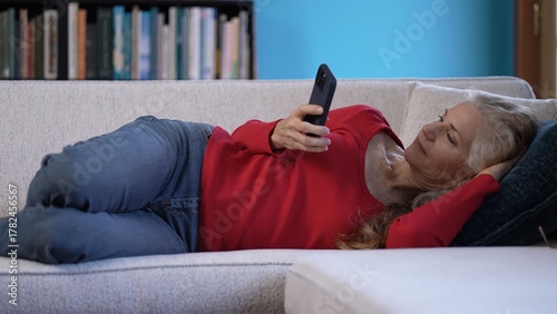 A mature woman lounges on a sofa, dressed in a red top and jeans, focused on her smartphone in a well-lit living room with bright blue walls and book shelves in the background.