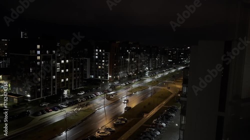 Street at night with moving car and illuminated residential buildings, wet asphalt with reflections,