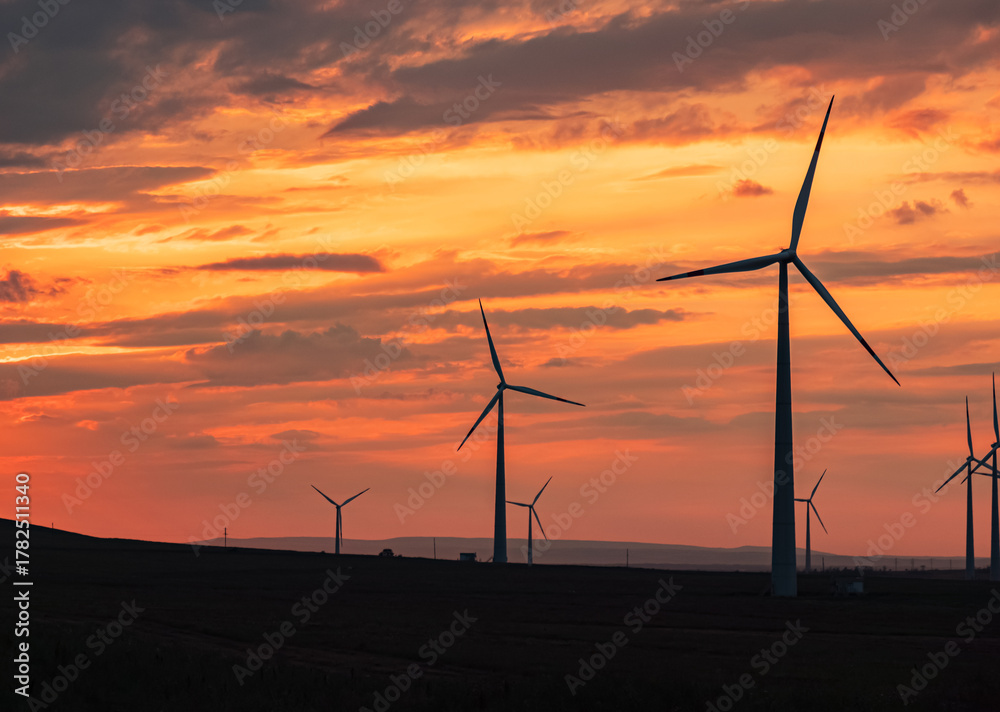 Obraz premium Multiple wind turbines silhouetted against a vibrant sunset sky. The turbines stand tall against the backdrop of orange and red clouds.
