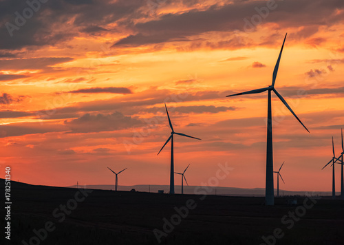 Multiple wind turbines silhouetted against a vibrant sunset sky. The turbines stand tall against the backdrop of orange and red clouds.