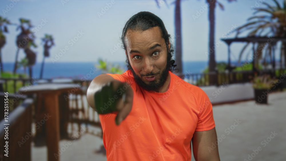 Fototapeta premium Man points finger to camera on building terrace, bare hand extended wearing orange shirt and beard; confidence invitation.