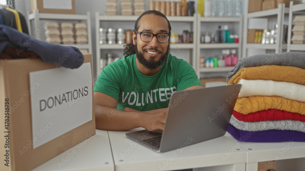 Naklejka premium Man in volunteer shirt types on a laptop beside a donations box and stacked sweaters in a building donation center; compassion.