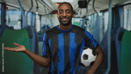 Slika na platnu Man holding soccer ball giving thumbs up inside bus on street, wearing blue black striped jersey and smiling; team pride