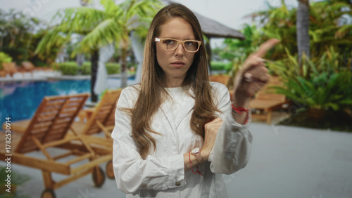 Woman scientist in lab coat with arms crossed and pointing finger at resort building poolside; serious professionalism.