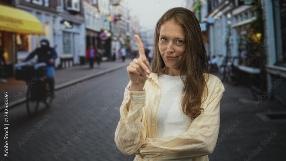 Fototapeta premium Woman with index finger raised and crossed arms on a cobblestone street in a striped shirt; playful warning.