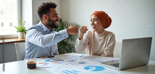 Man and woman give fist bump celebrating success at office desk with laptop and charts. Team members feel happy, showing partnership and good results. They work together effectively.