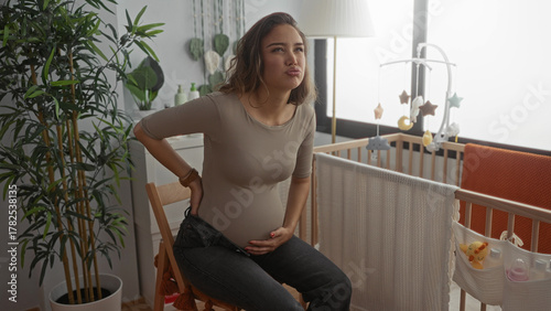 Pregnant woman sitting in nursery experiencing discomfort holding belly surrounded by crib and baby decor