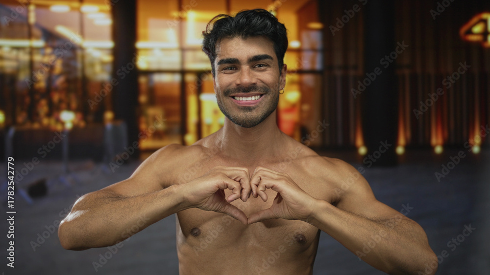 Fototapeta premium Young hispanic shirtless man forming heart shape with hands under street lights on busy urban street at dusk; love.