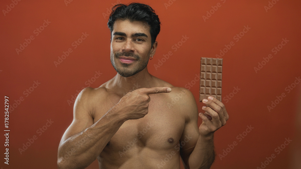 Fototapeta premium Muscular shirtless man points finger to a chocolate bar while smiling in an orange studio; temptation indulgence.