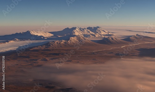 Majestic snow-capped mountains rise above a vast, arid landscape under a clear sky with a layer of fog at the base