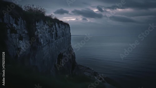 Dramatic cliff face overlooking a dark, moody sea under a cloudy twilight sky, rugged coastline and ocean expanse at dusk