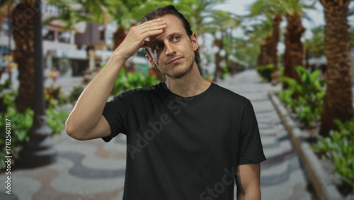 Young hispanic man with long hair and a black t shirt touching his forehead with one hand on a sunny street lined with palm trees and green plants; resignation.