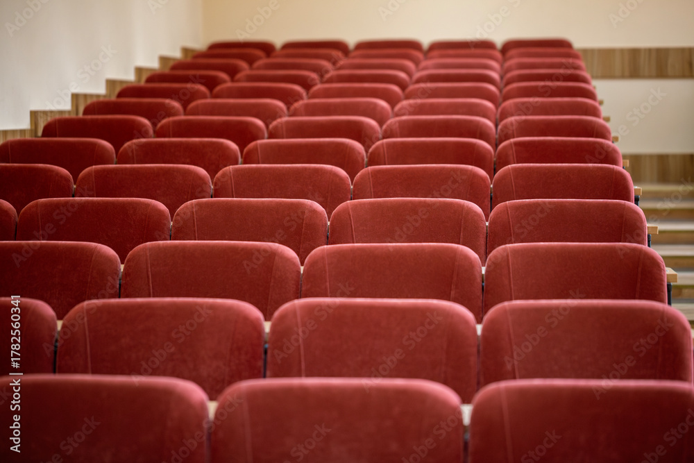 Fototapeta premium A close-up, front-on view of the empty maroon seats in a university lecture hall. The soft, upholstered chairs are arranged in neat rows, with wooden steps visible on the side.