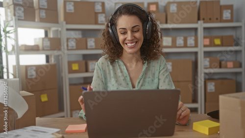 Woman wearing headphones smiling and gesturing with pen at laptop amid parcels on shelves in building; small business support joy.