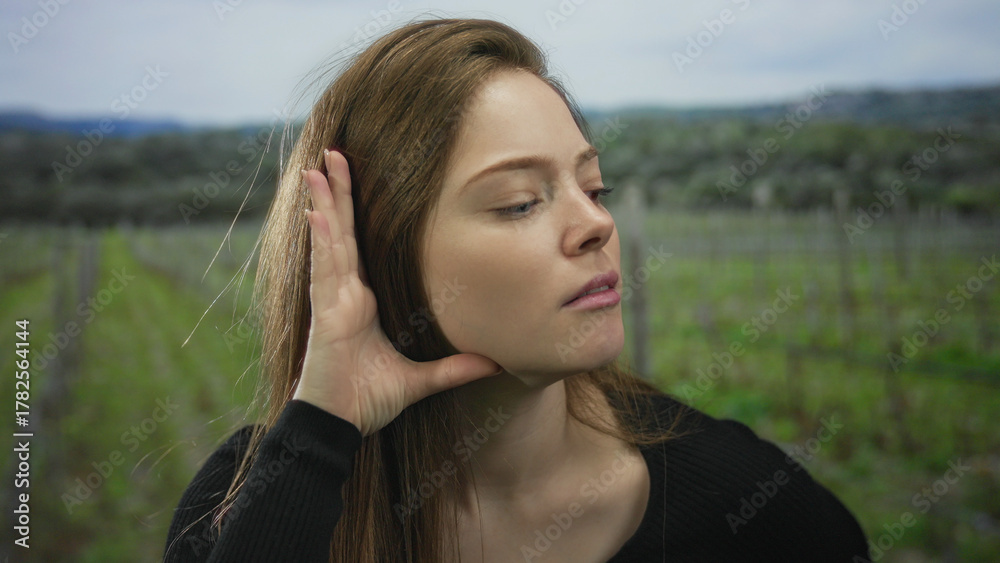 Fototapeta premium Woman cups ear with hand and closes eyes among green vineyard rows under cloudy sky in forest; curiosity.