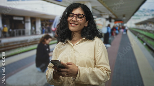 Fotografie Woman holding smartphone, hands typing, standing on a train platform inside a station building with rails visible; calm focus