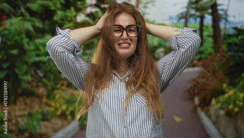 Foto Woman with glasses covers ears and holds head amid lush green forest pathway under sunlight; stress