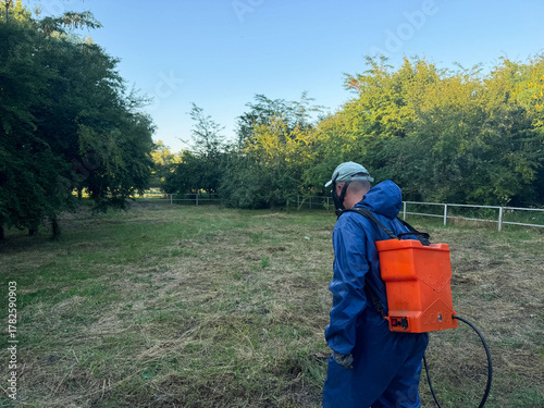 Man in protective gear sprays in a field surrounded by trees during early morning hours