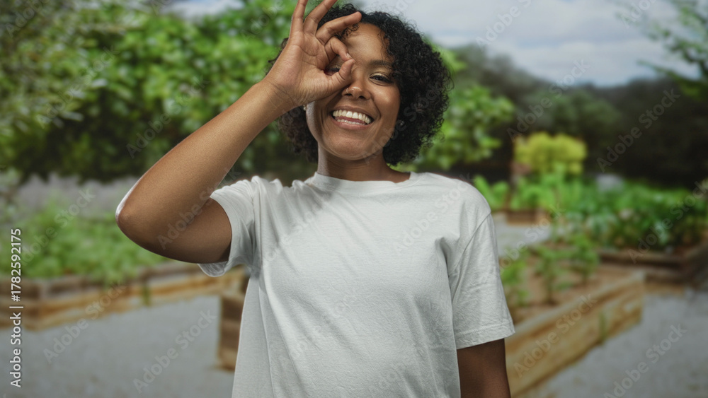 Obraz premium Woman makes ok sign with hand over eye in a green park with raised garden beds and foliage in background; approval.