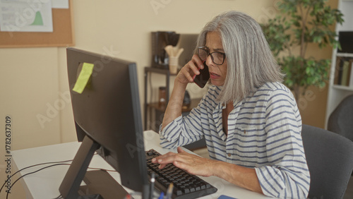 Valokuva Woman with grey hair and glasses holds phone to ear while typing on a keyboard at a desktop monitor in an office building; work concern