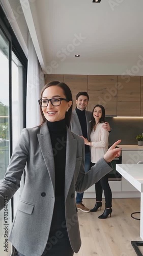 Vertical shot of a female real estate agent in the foreground, leading a happy young couple in the background who are viewing a modern apartment with an open-plan kitchen and large windows.