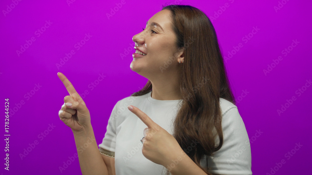 Obraz premium Hispanic young woman smiling and pointing both index fingers to the left in studio scene; excitement.