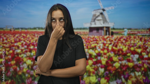 Young hispanic woman in black t shirt pinches nose while crossing arms among colorful tulip flowers near windmill building under blue sky; nuisance odor disgust.