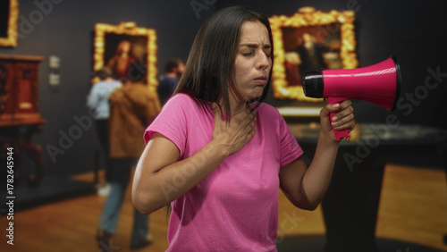 фотографія Young hispanic woman shouting into pink megaphone in an art gallery building while gesturing with hand to throat; urgent protest