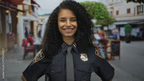 Young hispanic woman police officer in uniform smiling confidently on a lively street outdoors with people and cafes in the background.