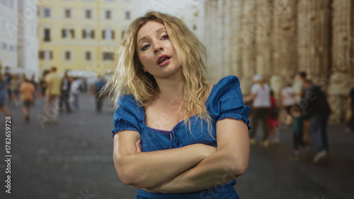 Photos Woman with arms crossed and windblown hair on street near historic building, blue dress visible; defiance