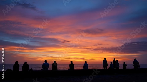 A silhouetted group of people contemplate a vividly colorful sunset over the ocean creating a serene evening scene