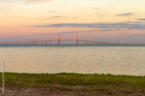 The Sunshine Skyway Bridge stretches gracefully across Tampa Bay at dawn, bathed in soft golden light as the new day begins. Gentle ripples on the calm water mirror the vibrant hues of the morning sky