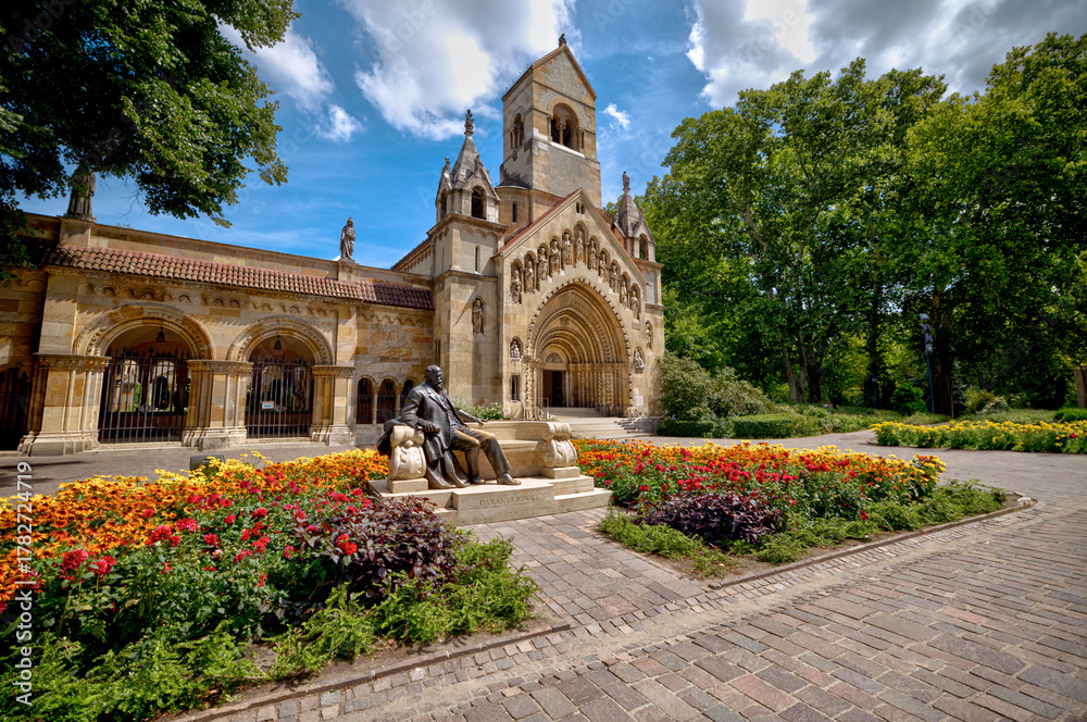 Naklejka premium Budapest, Hungary - July 27, 2025: The Romanesque-style Jak Chapel and the Anonymous statue at Vajdahunyad Castle in City Park, surrounded by colorful summer gardens 