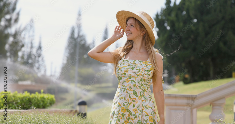 Fototapeta premium Smiling woman in summer dress and hat in sunny park, enjoying nature. Outdoor leisure, relaxed lifestyle, happiness concept.