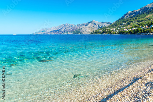 Fototapeta Naklejka Na Ścianę i Meble -  Beautiful pebble beach in Brela, Croatia with clear turquoise sea, calm waves, and mountains under a bright summer sky.