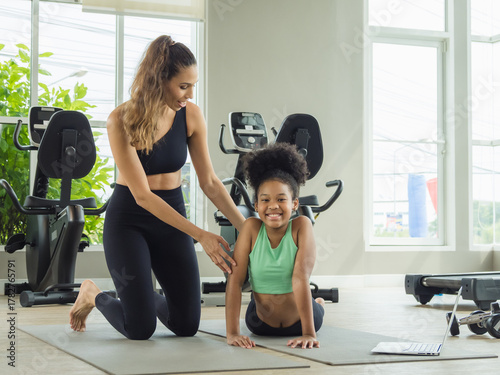 mother and daughter lying on exercise mats in a home gym, smiling while looking at a laptop screen. The scene highlights family bonding, fitness, and the use of technology for home workouts.