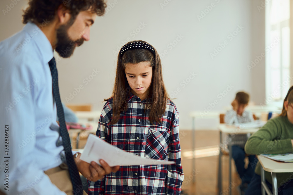 Obraz premium Sad elementary student talking to teacher while taking test results on a class in the classroom.