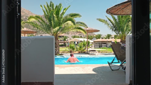 Man running out of hotel room and jumping into pool surrounded by palm trees on sunny day. Kos. Greece.