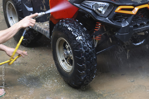 Man washing muddy red off road vehicle with water hose, showing satisfaction of cleaning after an adventure. Close up of hands cleaning dirty tire with high pressure spray