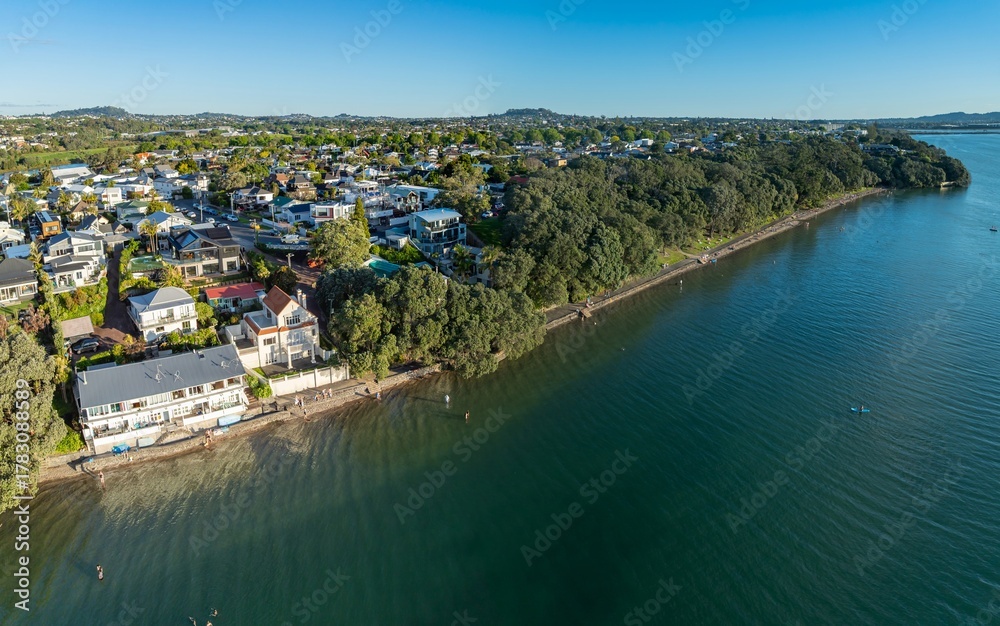 Fototapeta premium Aerial view of Point Chev, Auckland, New Zealand, showing people enjoying the beach and water. Some are swimming, walking along the shore, and paddle boarding on a sunny day.