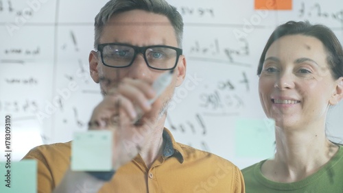 Two business people colleagues are analyzing notes on a transparent whiteboard during a brainstorming session, working together to develop innovative solutions and strategies for their deal