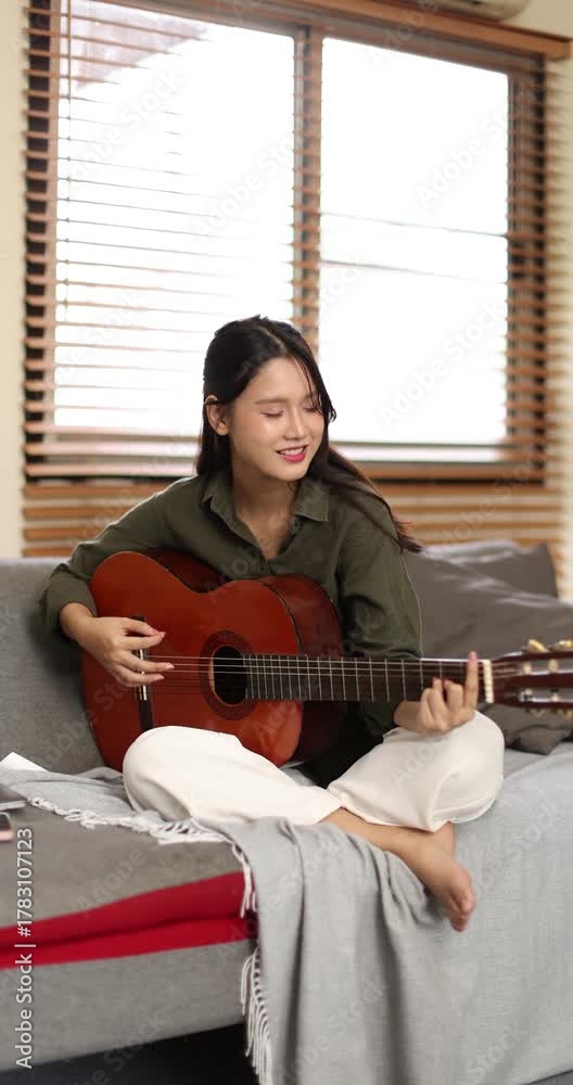 Asian young woman playing guiter sitting on coach alone in apartment