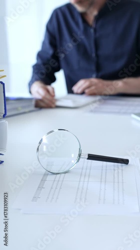Magnifying glass resting on a financial report, with a businessman focused on a laptop in the blurred background, emphasizing financial analysis and due diligence. Audit and taxes theme in business