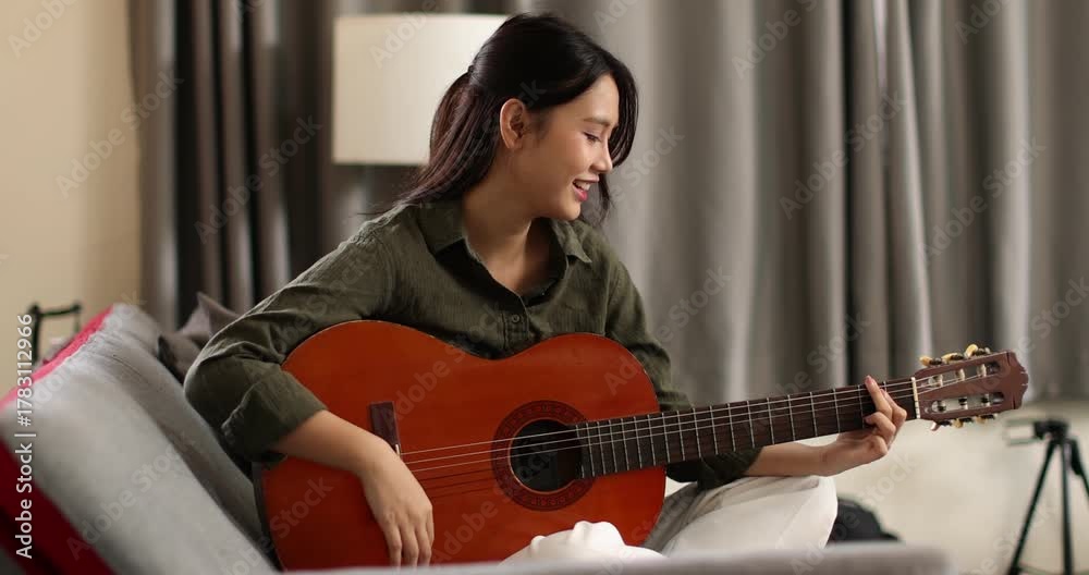 Asian young woman playing guiter sitting on coach alone in apartment