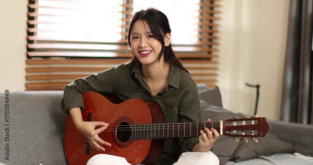 Asian young woman playing guiter sitting on coach alone in apartment
