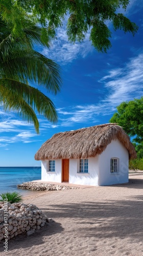 White Tropical Thatched Bungalow With Wooden Door Beside The Ocean Under A Clear Blue Sky With Lush Green Trees Framing The Scene