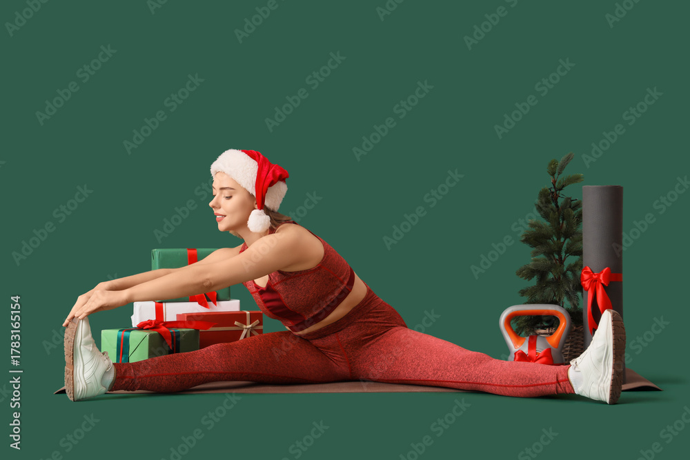 Fototapeta premium Sporty young woman with Christmas gift boxes, kettlebell and yoga mat exercising on green background